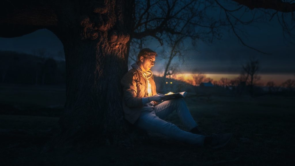 A man sitting with his back against a tree at night reading an open book that is glowing and illuminating his face.