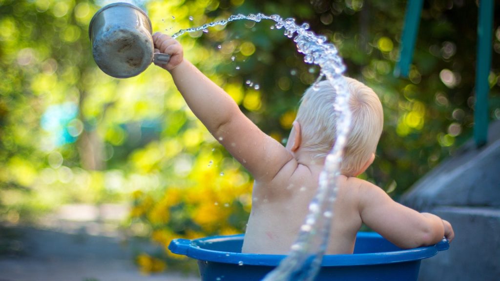 A toddler having a bath in a bucket outdoors swinging and spraying a tin cup of water in the air.