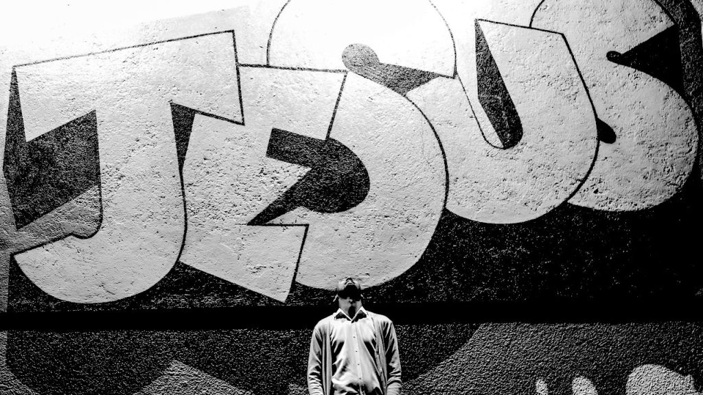 A man looking up at a giant graffiti version of the word Jesus.