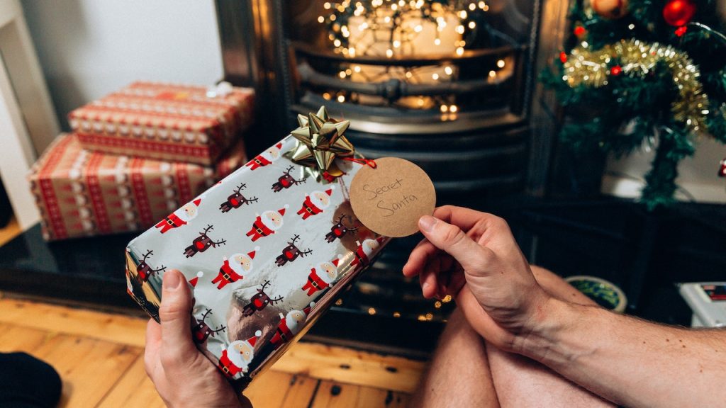 A person holding a gift-wrapped present at Christmastime in front of a fireplace.