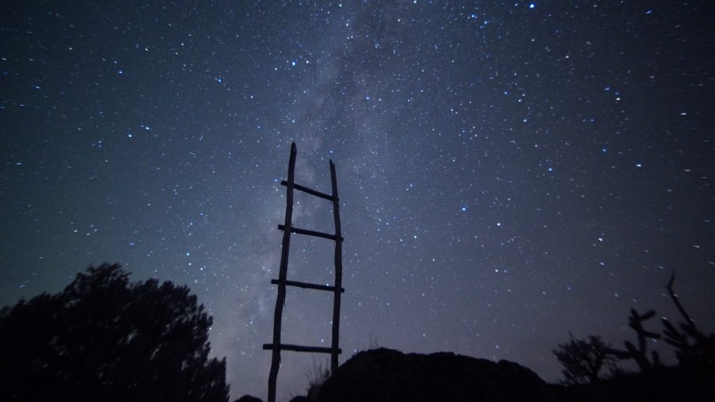 A silhouette of a ladder reaching up to a starlit sky at night.