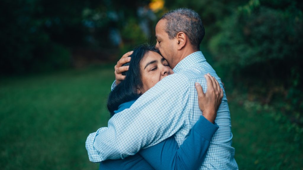 A couple standing in a garden at dusk tightly hugging one another.