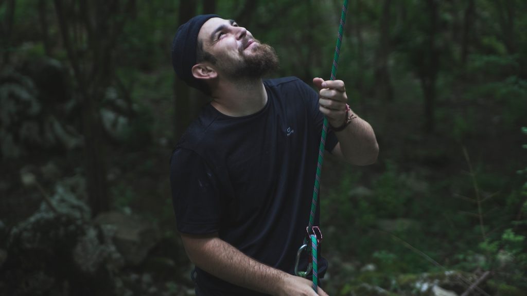 A man wearing a beanie, standing in a forest, looking up as he holds a climbing rope or belay line.