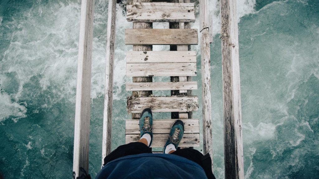 A person standing looking down at their feet as they cross a rickety wooden bridge over a raging river.