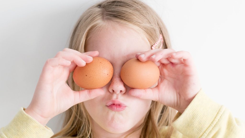 A young girl holding two hen eggs over her eyes and pulling a silly face.