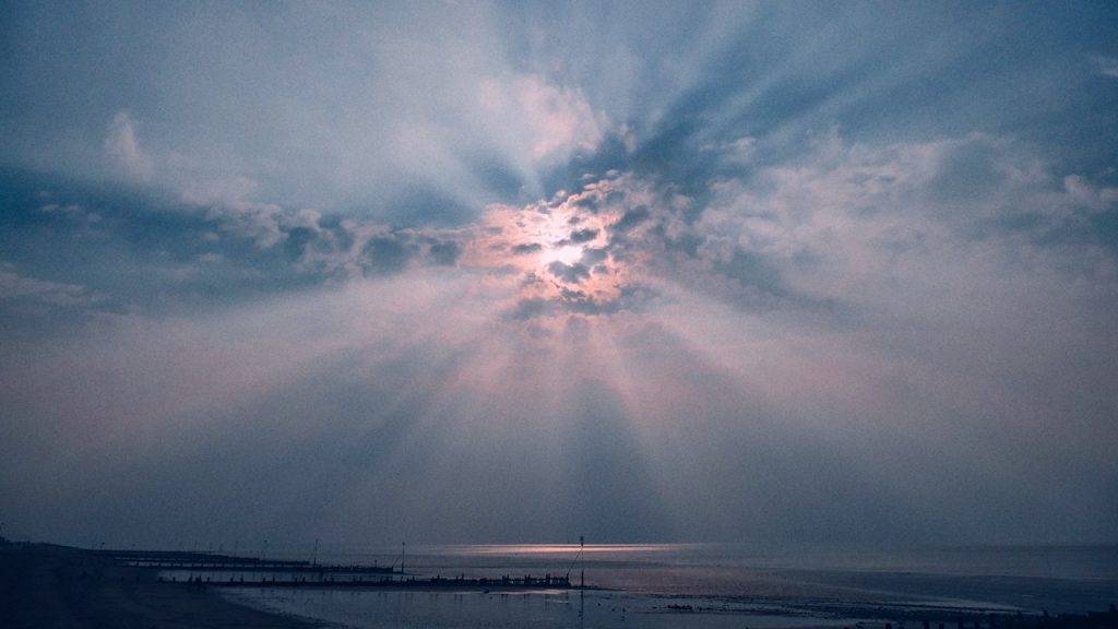 The sun bursting through a cloudy sky over the sea and sending rays off in all directions.