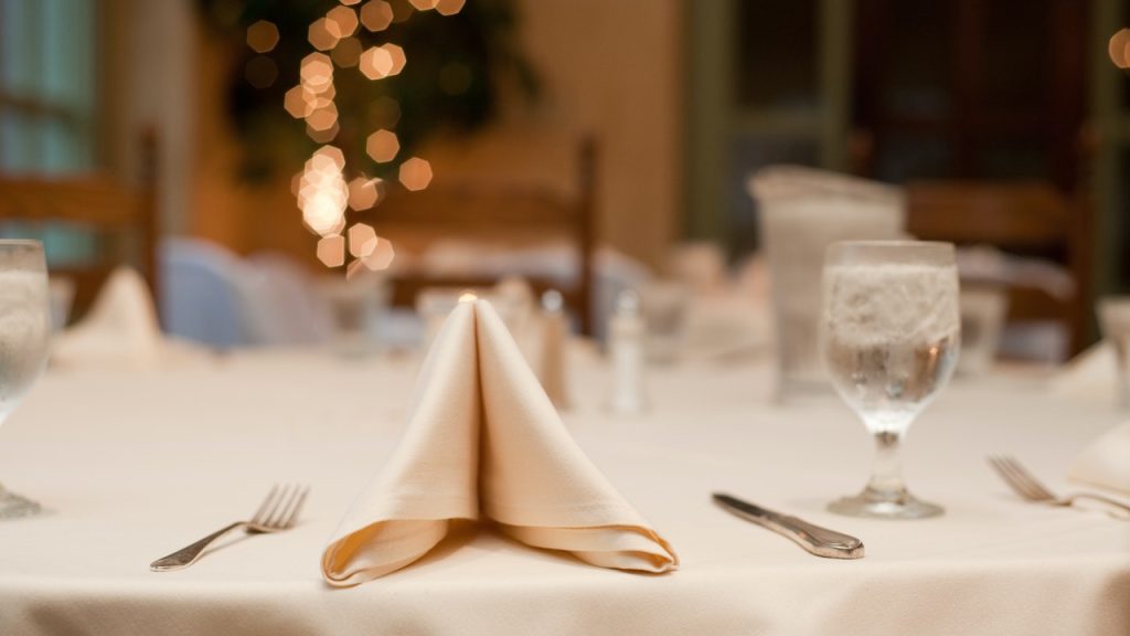 A closeup of a beautiful place setting on a table with silver cutlery, folded cream napkin, and crystal glass.