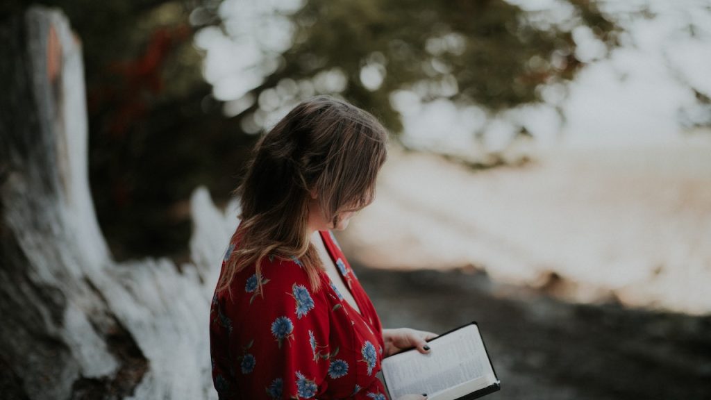 A women standing, looking down at an open Bible outdoors in a wild place.