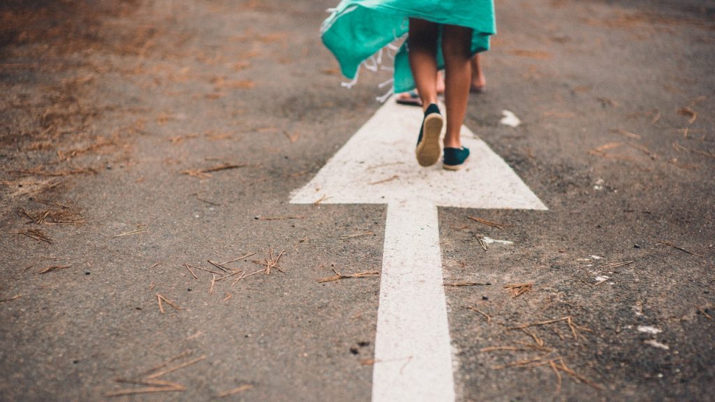 A person walking down a leaf-strewn road standing at the end of a large painted arrow on the road.