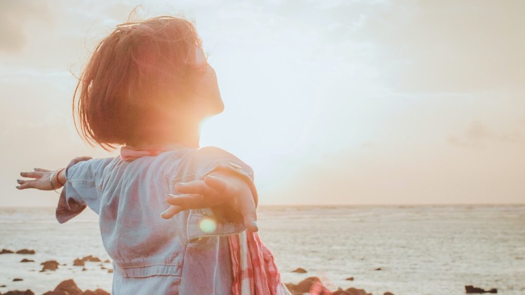 A woman standing on a sea shore with her head raised and arms stretched out bathing in the sunlight.