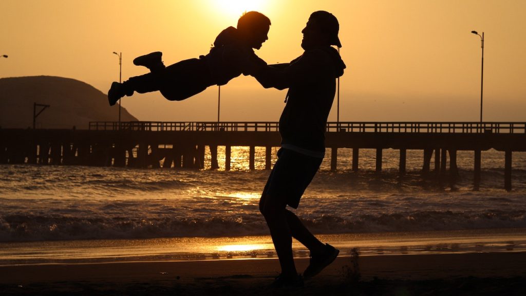 A silhouette of a man lifting a boy into the air as they stand on a beach at sunset.