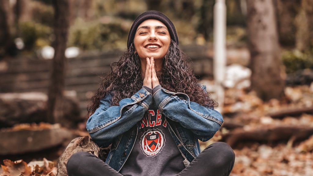 A woman sitting cross-legged on a forest floor, smiling, with her eyes closed and her hands pushed together in prayer.