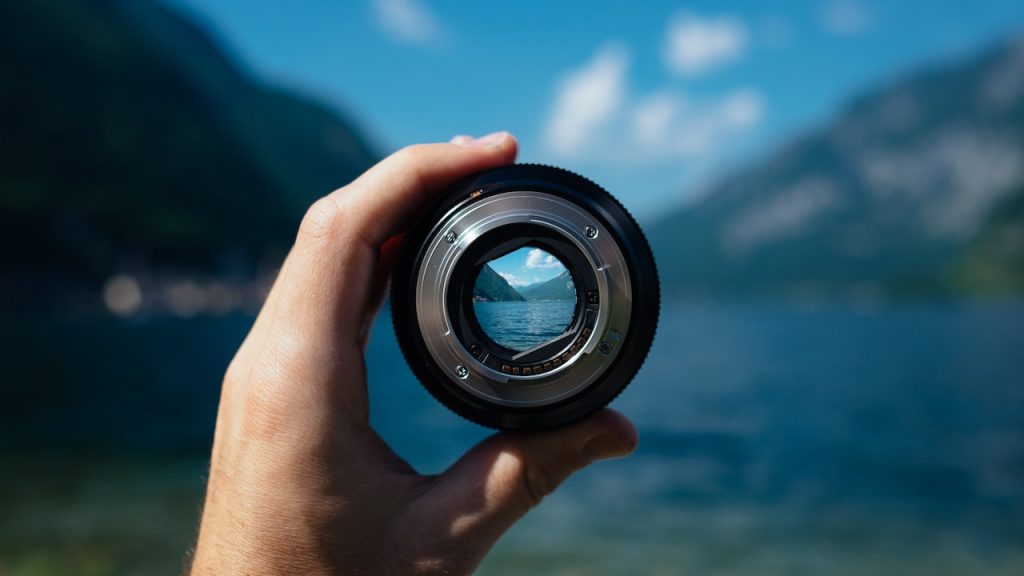 A hand holding a camera lens up so you can see through it so the whole vista of a fjord is in focus in the circle in the middle of the lens.