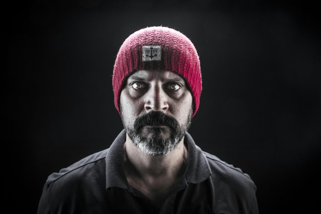 A bearded man wearing a red beanie hat frowns and glares directly at the camera looking foreboding and threatening.