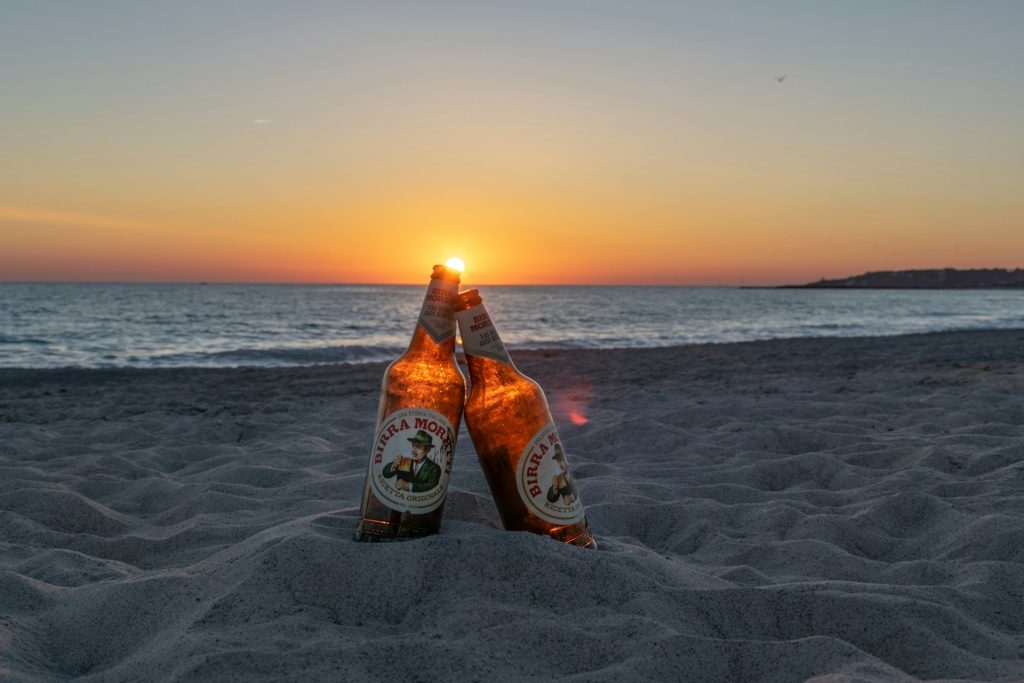 Two bottles of beer leaning against each other on a sandy beach at sunset.