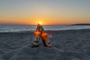 Two bottles of beer leaning against each other on a sandy beach at sunset.