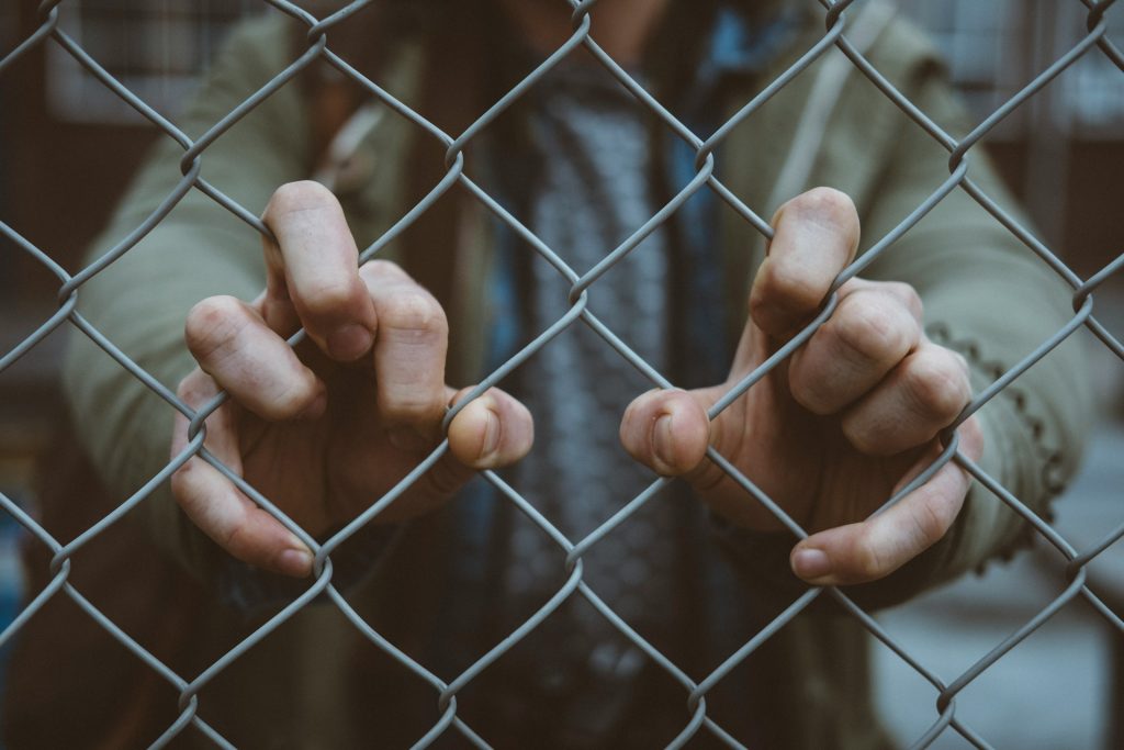 A person stands on the other side of a metal wire fence, their hands and fingers holding onto the wire.