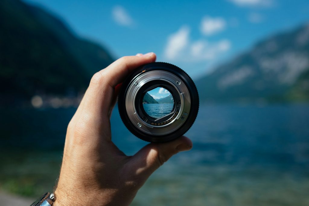 A camera lens is held up to eye level against a spectacular mountain and lake backdrop. You can see through the lens and the backdrop in the circular lens is in focus.