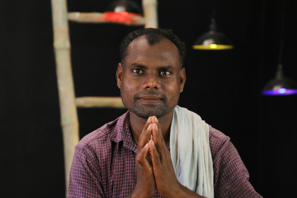 An Indian man with a serene expression in his face, sitting in a dark drama studio holding his hands together in prayer.