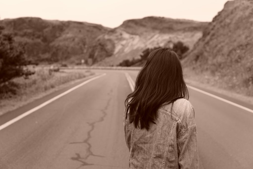 A woman standing in a road in the mountains looking away down the road into the distance.