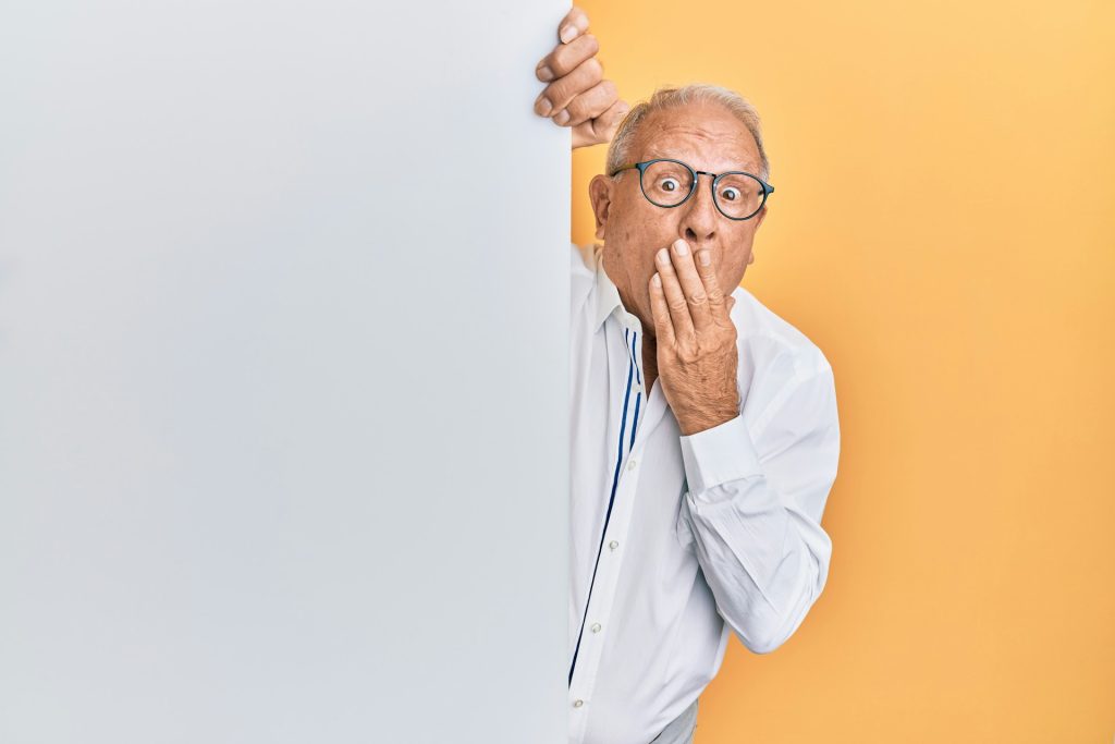 A man with his hand over his mouth looking shocked, leaning around the corner of a wall and looking at the camera.