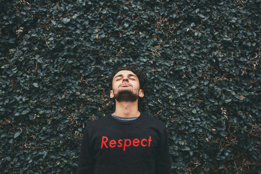 A man looking upwards leaning against a thick hedge. He is wearing a shirt with the word Respect in large letters written on it.