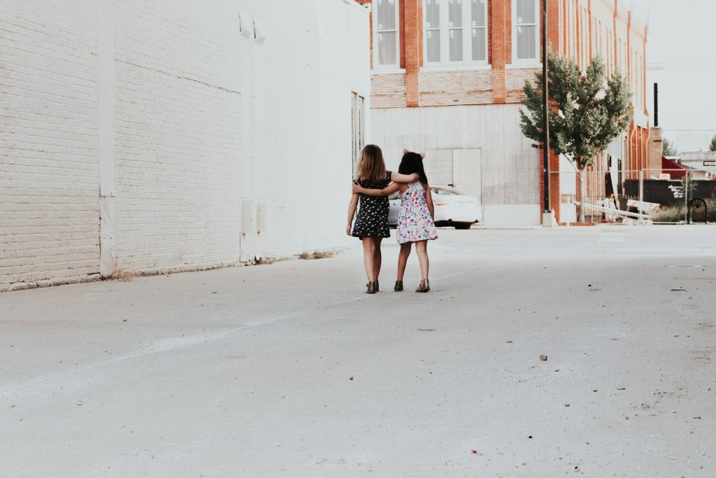 Two young girls with their arms around each other walking in an empty urban street.