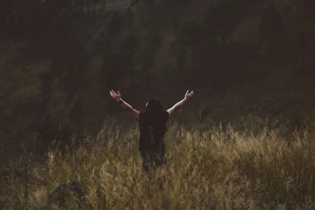 A women standing in a field of long grass in a forest with her arms outstretched towards heaven.