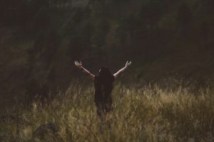 A women standing in a field of long grass in a forest with her arms outstretched towards heaven.