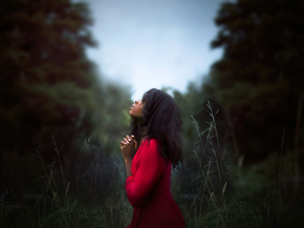 A woman, wearing a red dress, stands alone in a green forest holding up her hands in prayer.
