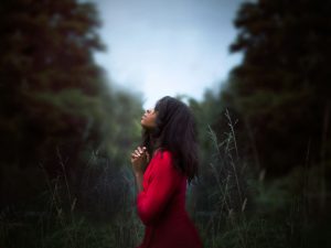 A woman, wearing a red dress, stands alone in a green forest holding up her hands in prayer.