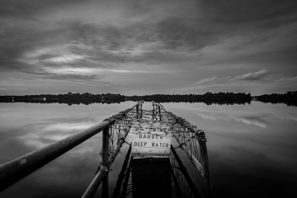 A jetty leading out into a lake is closed off with a sign saying "Danger. For Your Safety. Deep Water."