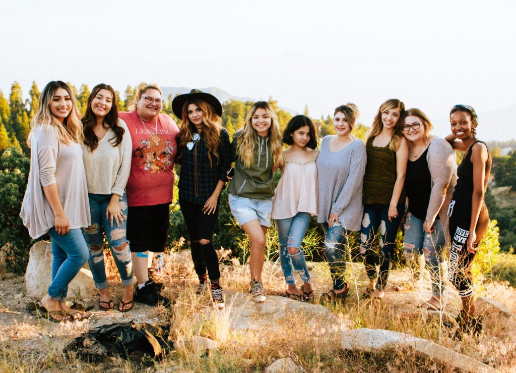 A group of diverse friends all stand in a line with their arms round each other.