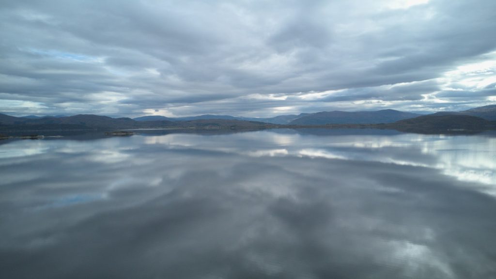 A flat calm lake with the clouds in the sky reflected in the water.