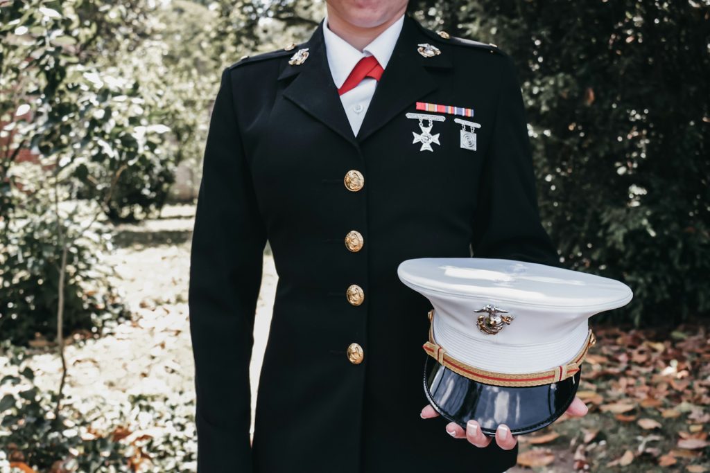 A female marine stands in smart uniform, holding her hat against a woodland backdrop.