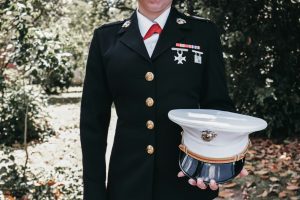 A female marine stands in smart uniform, holding her hat against a woodland backdrop.