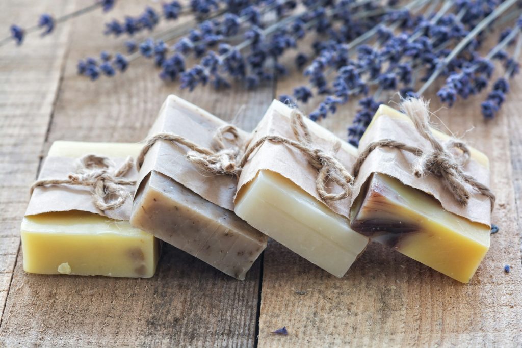 Four bars of beautiful hand-made lavender soap lie on a counter with som freshly picked lavender flowers.