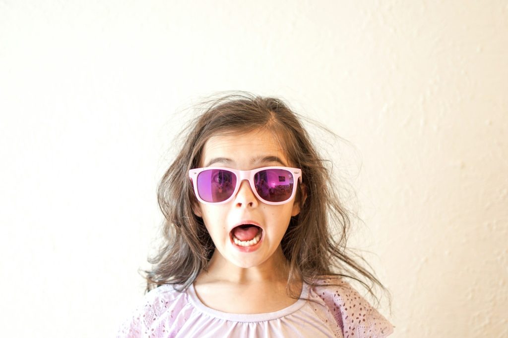 A young girl, wearing purple oversized sunglasses stands with her eyebrows raised and her mouth wide open. A look of surprise!