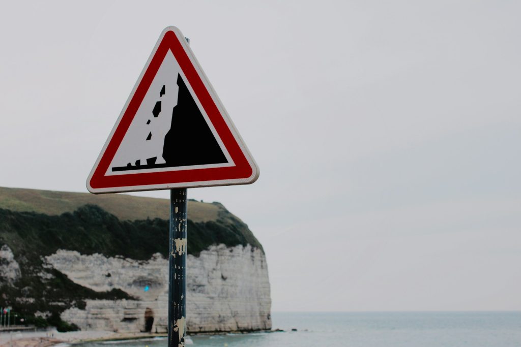 A red triangular warning sign with an image of black rocks falling from a cliff stands on a post in front of a coastal white cliff.