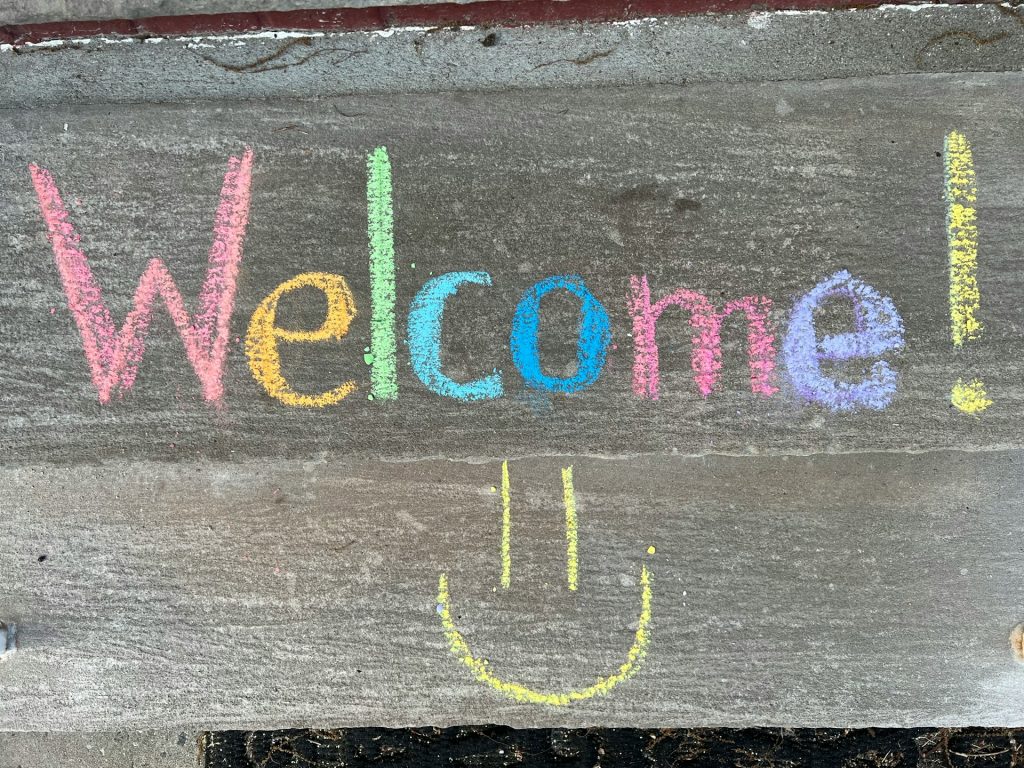 The word welcome has been chalked onto a wooden board with each letter a different colour. Underneath the word has been drawn a smiley face.