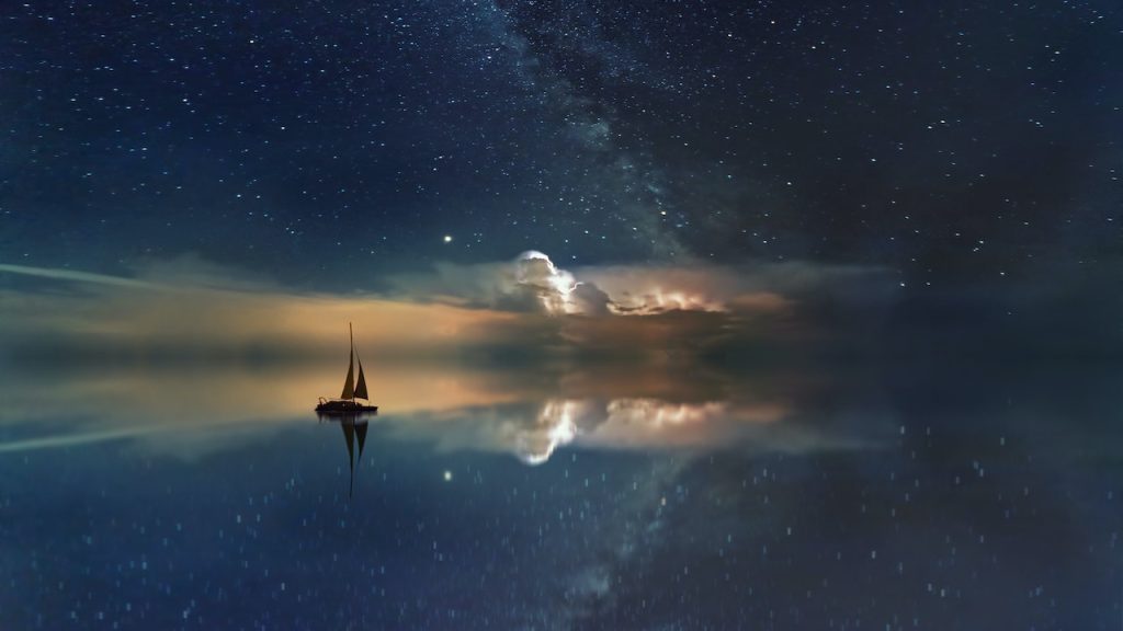 A silhouette of a sailing boat in a vast ocean with the starlit sky reflected on the calm water.