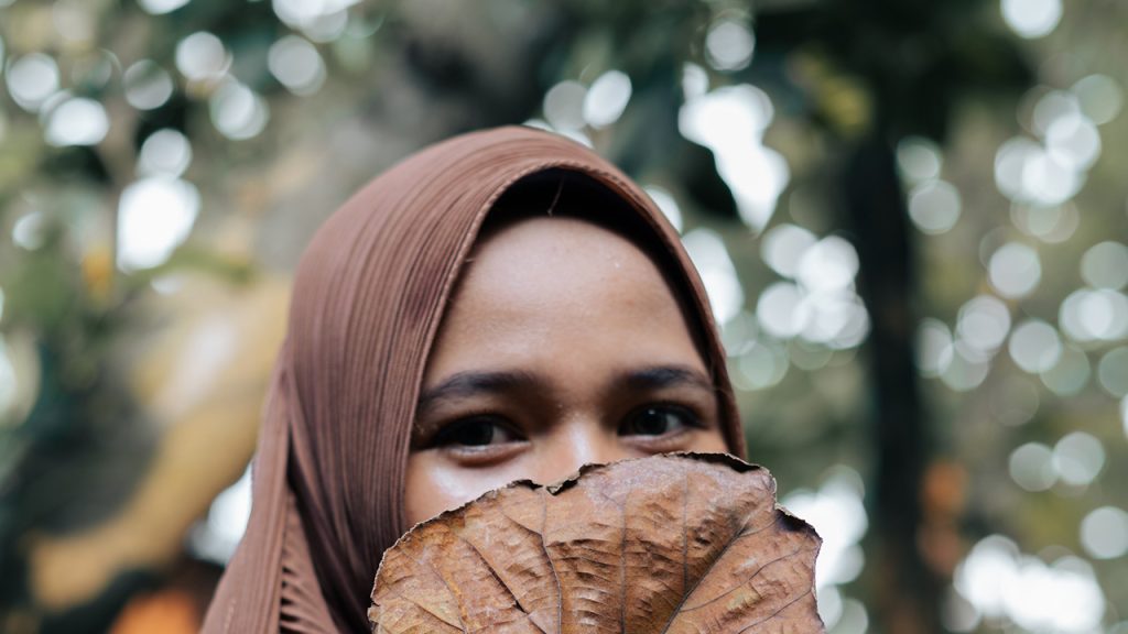 A woman with a head scarf covering her face with a large brown leaf so you can only see her smiling eyes.