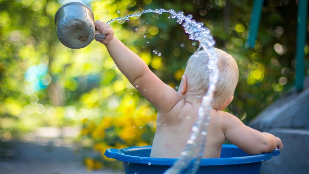 A bathing toddler sitting outside in a plastic bucket flinging water into the air from a tin cup.