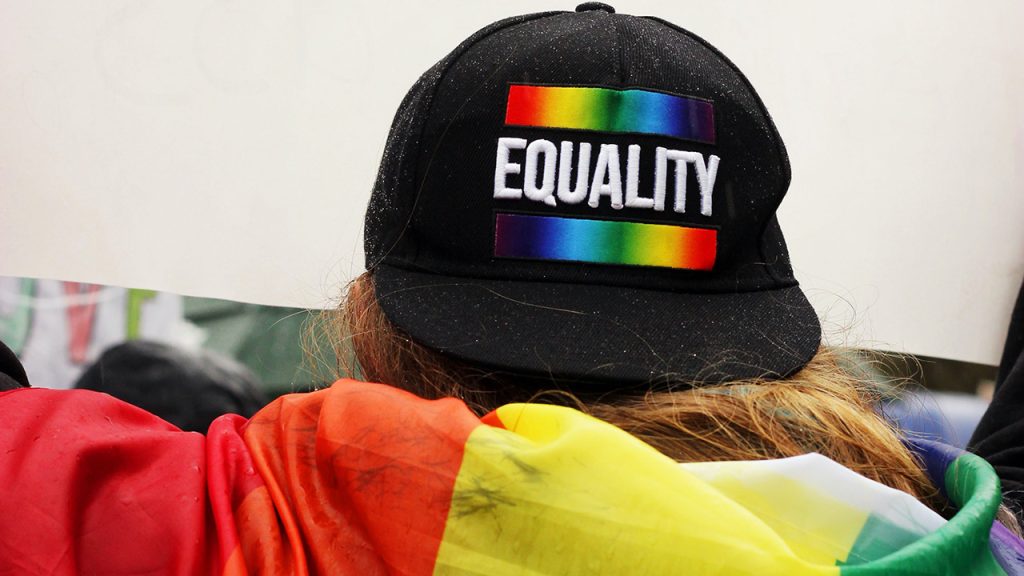 A person draped in a rainbow flag wearing a hat on backwards with the word EQUALITY written on it.