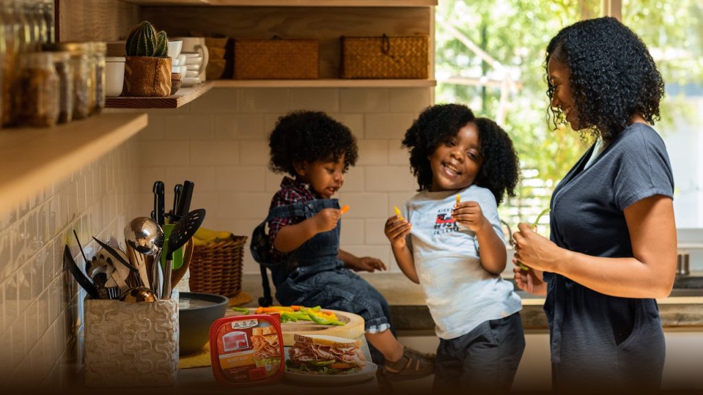 A woman and two children in a kitchen making and eating ham sandwiches.