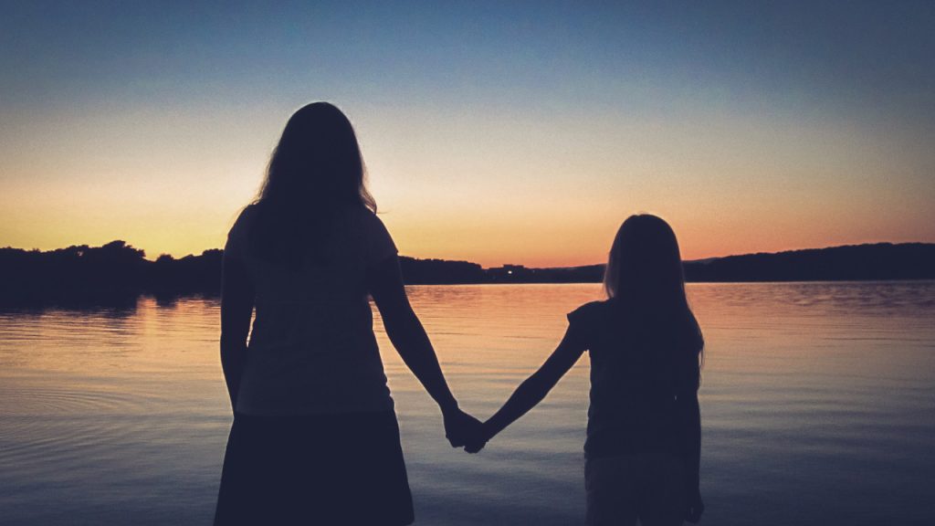 A silhouette of a young girl holding her mother's hand as they look out over a lake at Sunset.