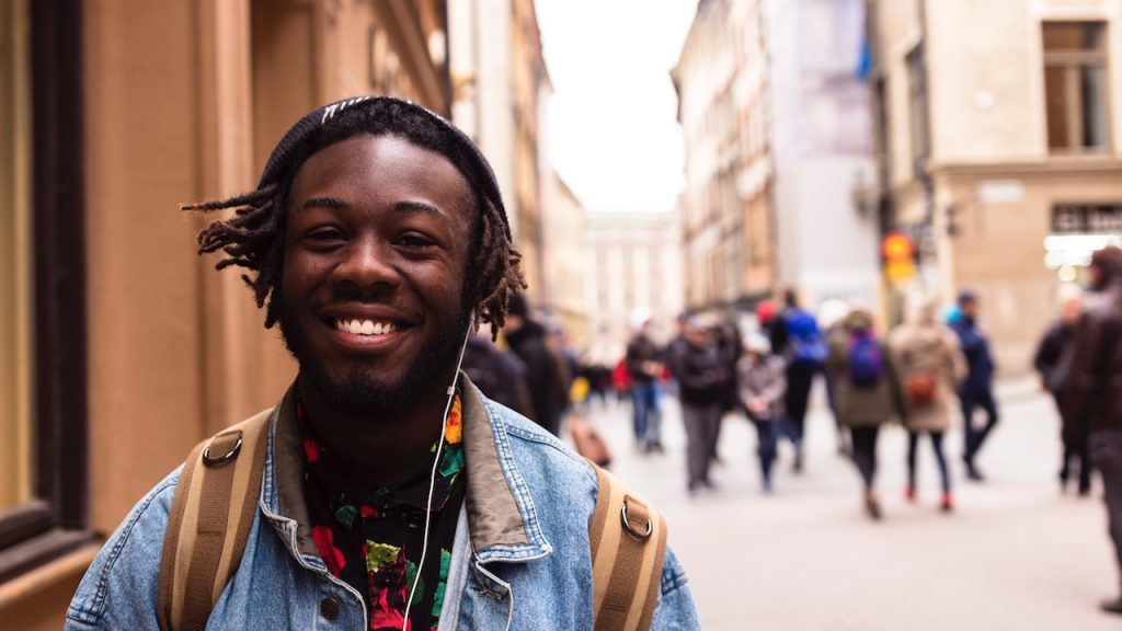 A young man wearing a beanie and headphones smiling and standing in an urban high street.