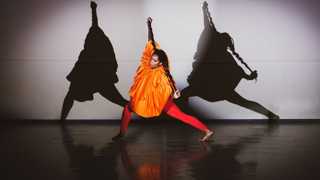 A woman in a dance studio dressed in bright colours with two shadows on the wall behind her.