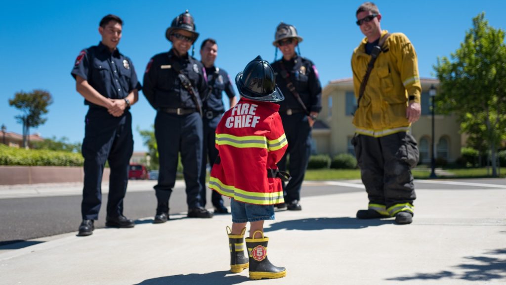 A kid in a fireman's uniform standing before several adult firefighters.