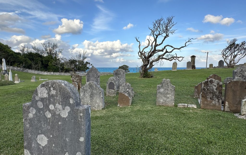 A cemetery with gravestones in the day time with old wisened trees and the sea in the background.